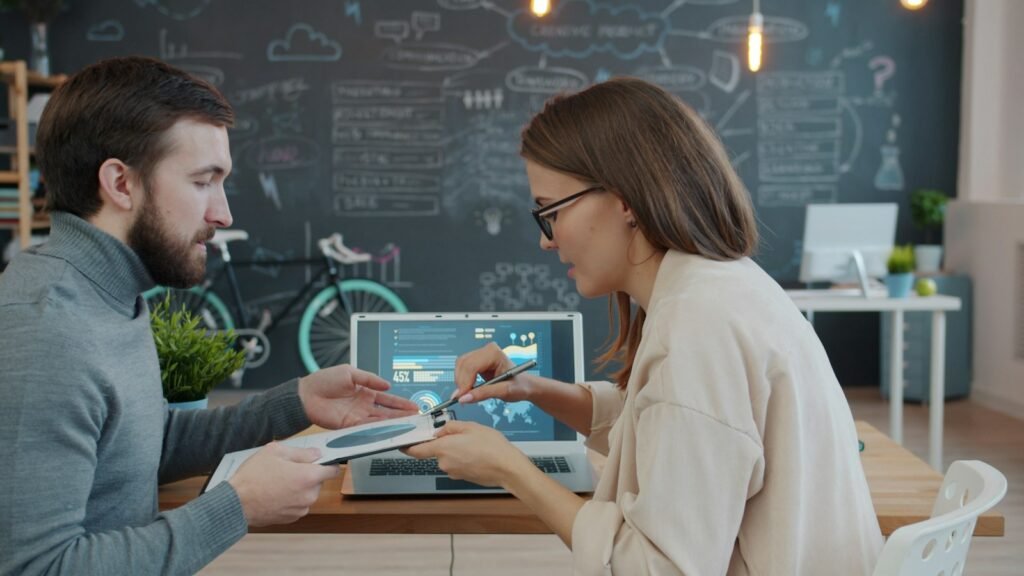 Two colleagues discussing information shown on a laptop and a clipboard.